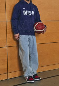 Sweat à capuche Nike violet avec logo NBA blanc, jean ample bleu clair et baskets noir/rouge. Tenant un ballon de basket contre un mur en bois.