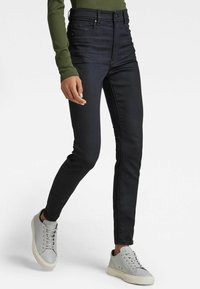 Černé skinny džíny s vysokým pasem a hladkým povrchem, s předním zipem a zapínáním na knoflík, doplněné o světle šedé tenisky.