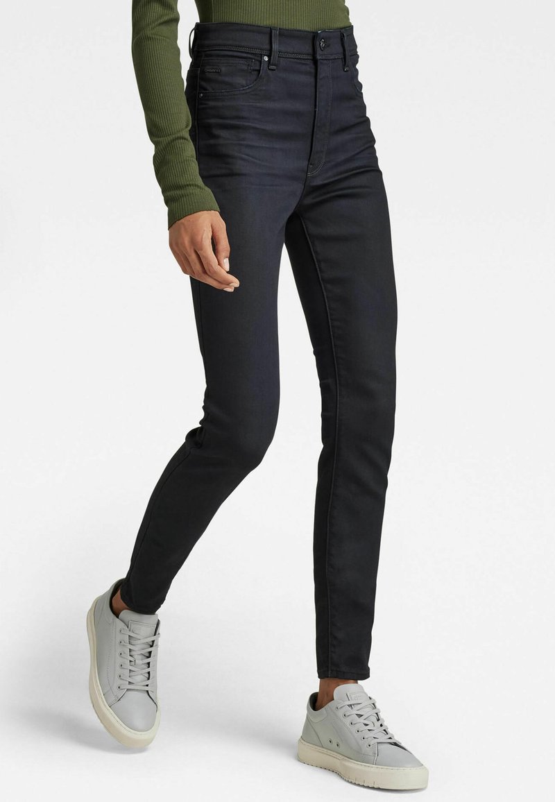 Černé skinny džíny s vysokým pasem a hladkým povrchem, s předním zipem a zapínáním na knoflík, doplněné o světle šedé tenisky.