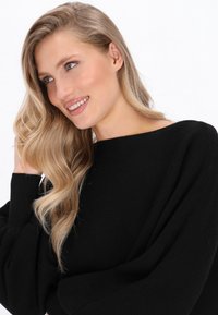 Schwarzer gerippter Pullover mit weitem Ausschnitt und voluminösen Ärmeln. Das Model hat lange wellige Haare und einen hellen Hintergrund.