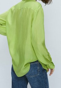 Green sheer long-sleeve shirt with a relaxed fit, featuring a rounded hem and slightly textured fabric, worn over blue jeans.