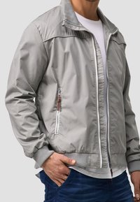 Man in een lichtgrijze zip-up jas over een wit T-shirt en blauwe spijkerbroek, met hand in zijn zak, tegen een effen lichte achtergrond.
