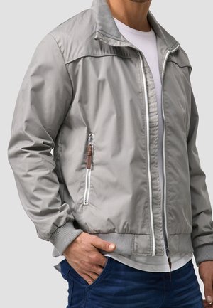 Man wearing light gray zip-up jacket over white t-shirt and blue jeans with hand in pocket, against plain light background.