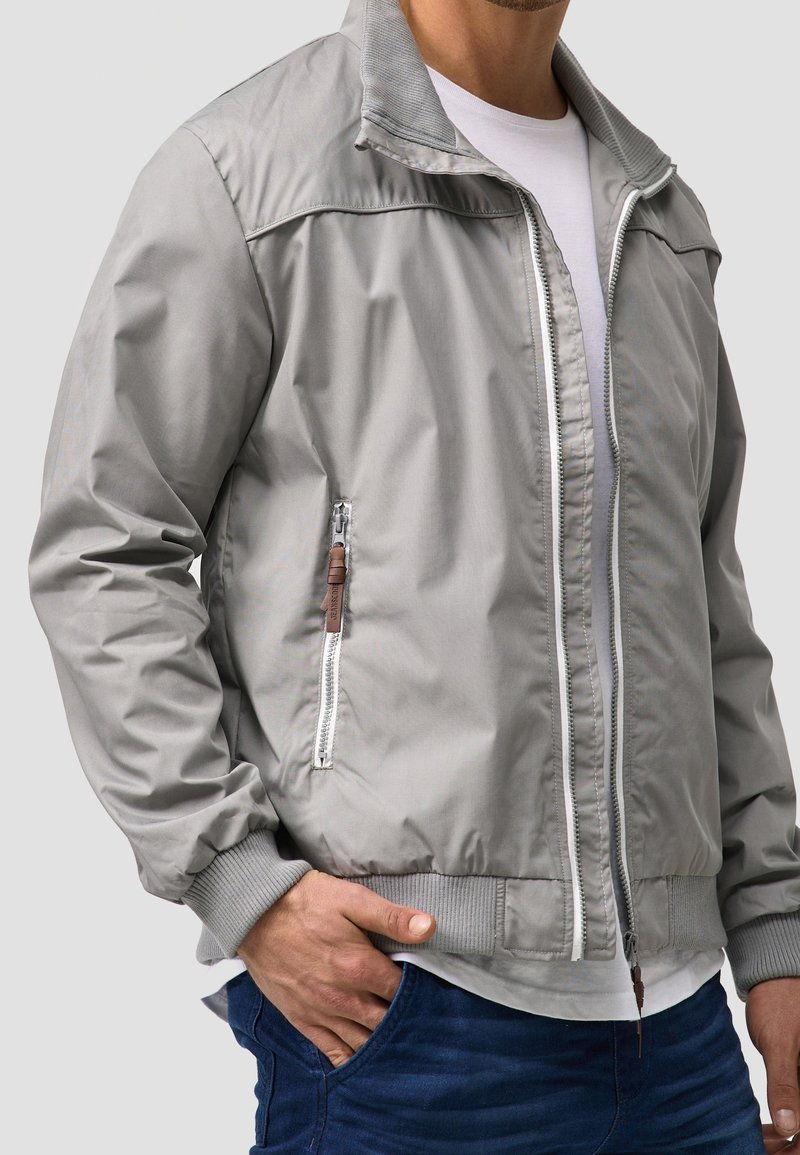 Man in een lichtgrijze zip-up jas over een wit T-shirt en blauwe spijkerbroek, met hand in zijn zak, tegen een effen lichte achtergrond.