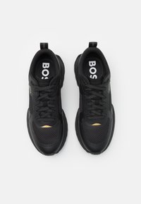 Zwarte sportieve schoenen met een bovenwerk van mesh, een gevoerde kraag en een vetersluiting. Gouden accenten nabij de neus. Rubberen zool voor grip.
