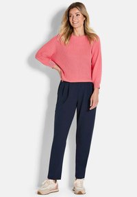 Femme aux longs cheveux blonds portant un pull côtelé corail, un pantalon plissé bleu marine et des baskets beiges, se tenant avec une main sur la hanche.