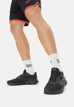 Pies con zapatillas deportivas negras Reebok y calcetines blancos Reebok, mostrados con piernas dobladas, llevando pantalones cortos negros con ribete rojo sobre un fondo blanco.