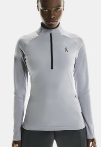Helles graues Langarm-Halbzip-Shirt mit strukturiertem Finish, das einen hohen Kragen und ein kleines Logo auf der linken Seite hat.