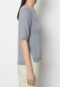 Kobieta w jasnoszarym swetrze z krótkim rękawem i kremowych spodniach, stojąca bokiem na jednolitym białym tle.