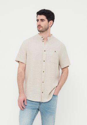 Mann steht, trägt ein beige kurzärmeliges Hemd mit Mandarinkragen und hellblaue Jeans, schaut zur Seite.