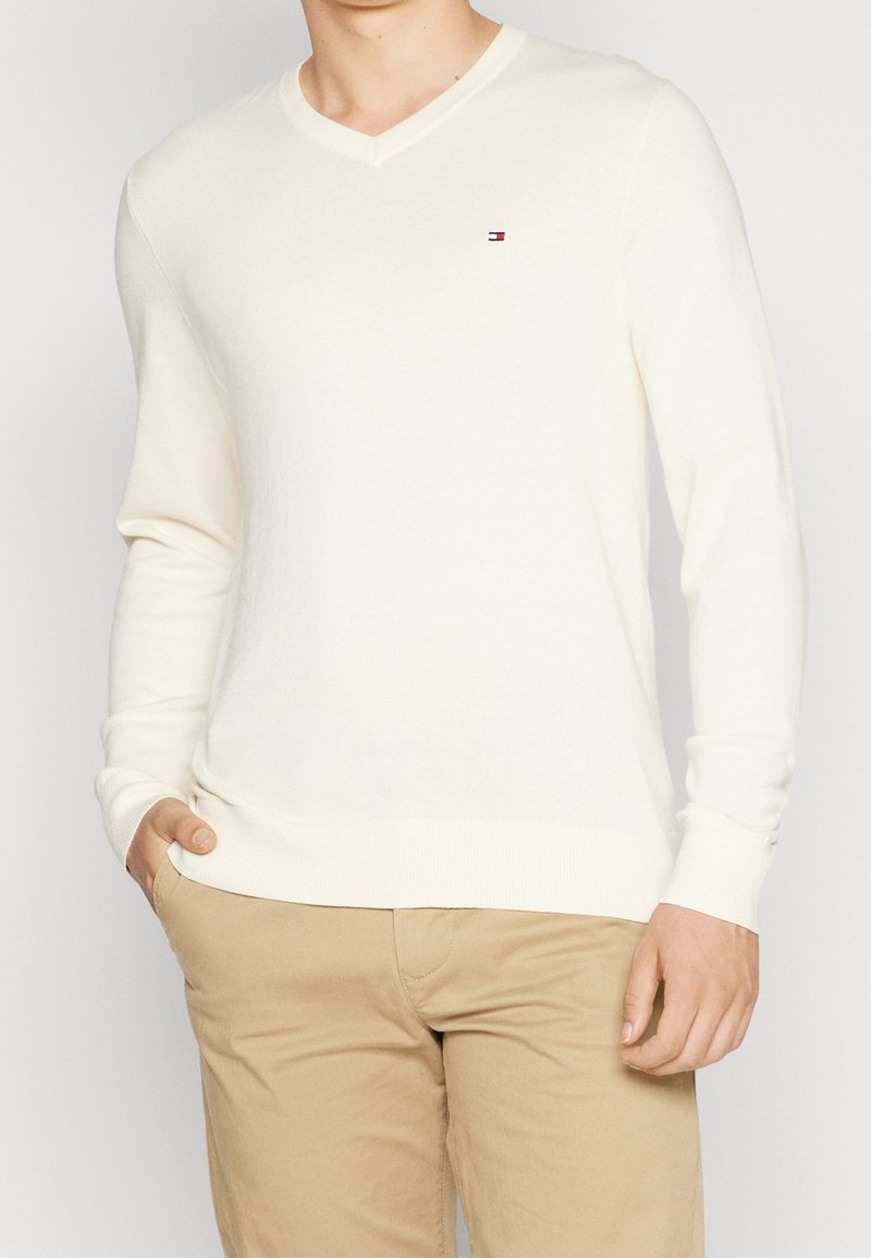 Uomo che indossa un maglione bianco a V con un piccolo logo rosso, bianco e blu sul petto e pantaloni beige, con una mano in tasca.