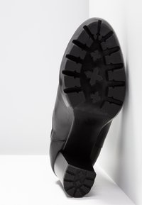 Black shoe sole with textured rubber, featuring deep treads and a wide block heel. Side view shows smooth leather upper with minimal stitching.