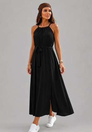 Femme portant une robe maxi noire sans manches avec une ceinture à la taille, un foulard marron, des bijoux dorés et des baskets blanches sur un fond gris uni.