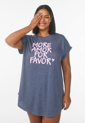 Smilende kvinde med mellemlangt hår dækker det ene øje med hånden, iført en løs blå kjole med teksten "More Amor Por Favor" i lyserøde bogstaver.