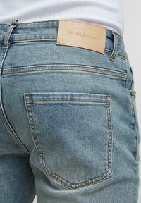 Primo piano di una tasca posteriore di jeans in denim azzurro chiaro e di una etichetta in pelle marrone sul waistband, indossati con una maglietta bianca a coste.