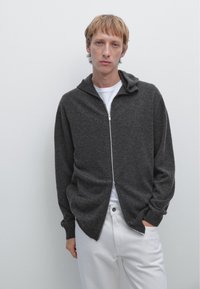 Grå zip-up hoodie med en strukturerad tyg, har en huva, långa ärmar och en avslappnad passform. Bärs över en vit T-shirt och matchas med vita byxor.