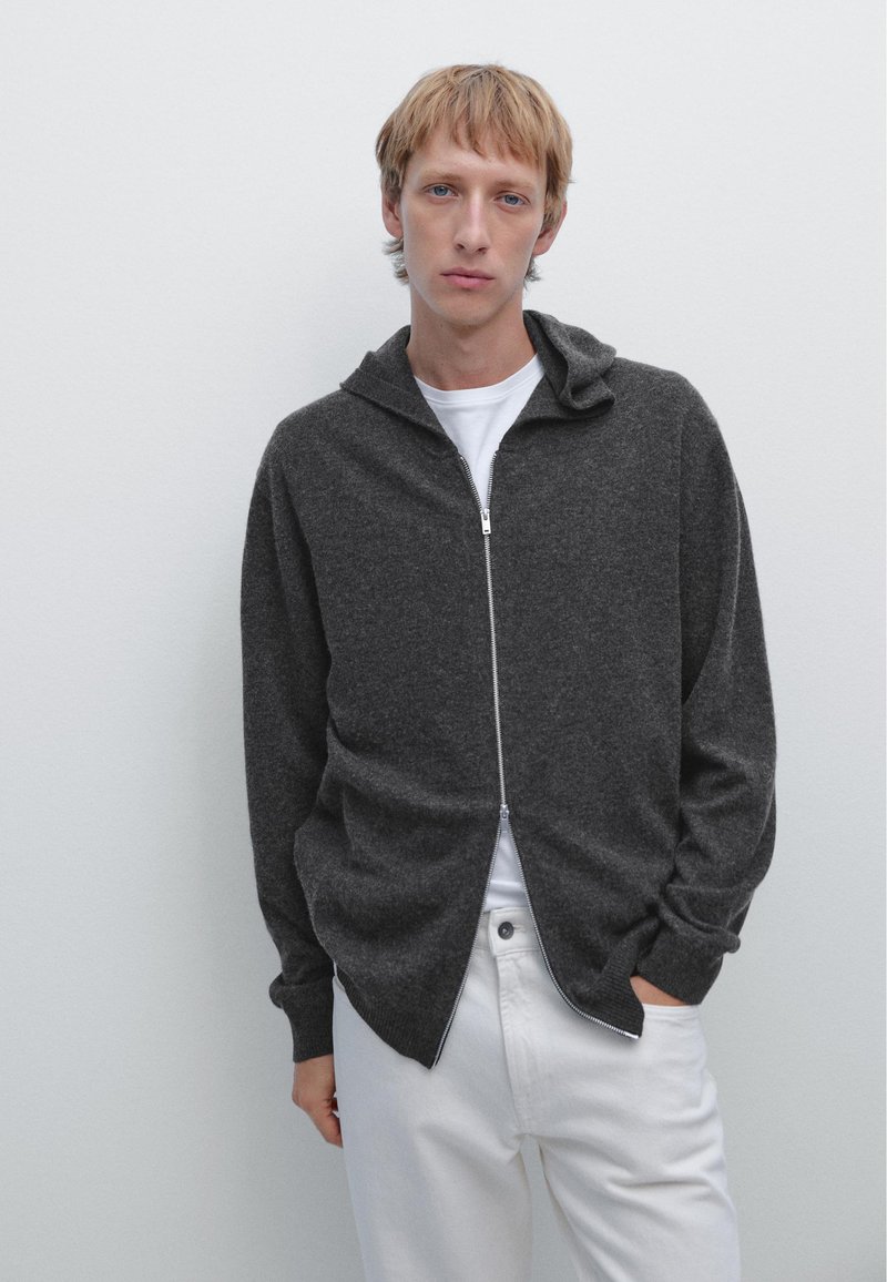 Grå zip-up hoodie med en strukturerad tyg, har en huva, långa ärmar och en avslappnad passform. Bärs över en vit T-shirt och matchas med vita byxor.