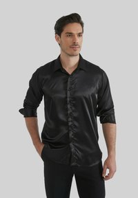 Chemise en satin noir avec un col classique, un devant à boutons et des manches retroussées, présentant une texture lisse et brillante.