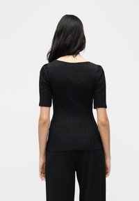 Γυναίκα με μακριά μαύρα μαλλιά που φοράει εφαρμοστό μαύρο κοντομάνικο ριμπ μπλουζάκι και φαρδιά μαύρη παντελόνα, στέκεται με την πλάτη στην κάμερα.