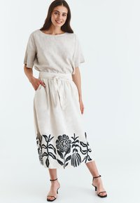 Linned to-delt outfit med en beige, kortærmet top og en lang nederdel med sorte blomster mønstre langs kanten. Tied taljedetalje.