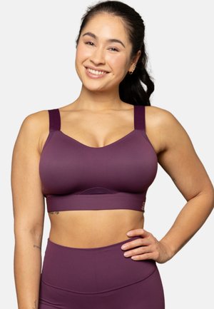 Smiling woman with dark hair in a ponytail wearing a purple sports bra and high-waisted leggings, hand resting on hip, against a white background.
