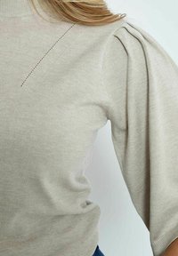 Beiger Strickpullover mit hohem Ausschnitt, subtilen diagonalen Nähten und gerafften Puffärmel. Weiche Textur mit einem leicht lockeren Schnitt.