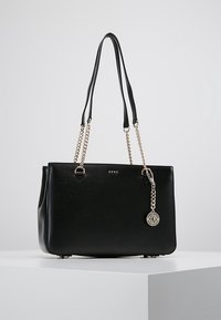 Sac à main rectangulaire noir avec des anses en chaîne dorée, logo "DKNY" et charme circulaire, exposé sur un piédestal blanc contre un fond gris.