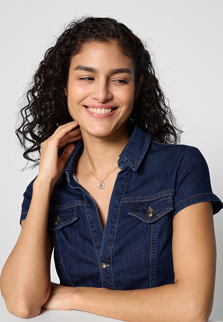 Dunkelblaue Denim-Bluse mit kurzen Ärmeln, Knopfdetails und Brusttaschen. Das Model trägt eine schlichte goldene Halskette und zeigt ein natürliches Lächeln.