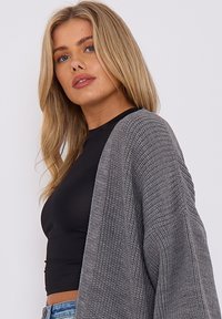 Grauer Strickcardigan mit lockerer Passform und gerippter Textur. Über einem schwarzen Crop-Top getragen, vor einem schlichten weißen Hintergrund ausgestellt.