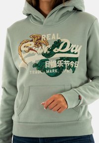 Lichtgroene hoodie met een frontzak, voorzien van geborduurde graphics van golven en een vogel in gouden en groene tinten.