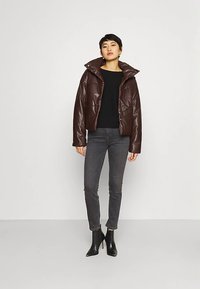 Hnědá krátká bunda z umělé kůže s vysokým límcem, zapínáním na patentky vpředu a vycpaným designem. Kombinována s černým topem a šedými úzkými džíny.