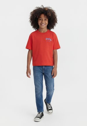 Enfant souriant aux cheveux bouclés marchant, portant un t-shirt rouge, un jean bleu et des baskets noires sur un fond blanc uni.