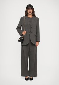 Donna in piedi con un pantalone grigio a righe sottili, che tiene una piccola borsa nera di paillettes con una catena dorata, indossando scarpe nere a punta.