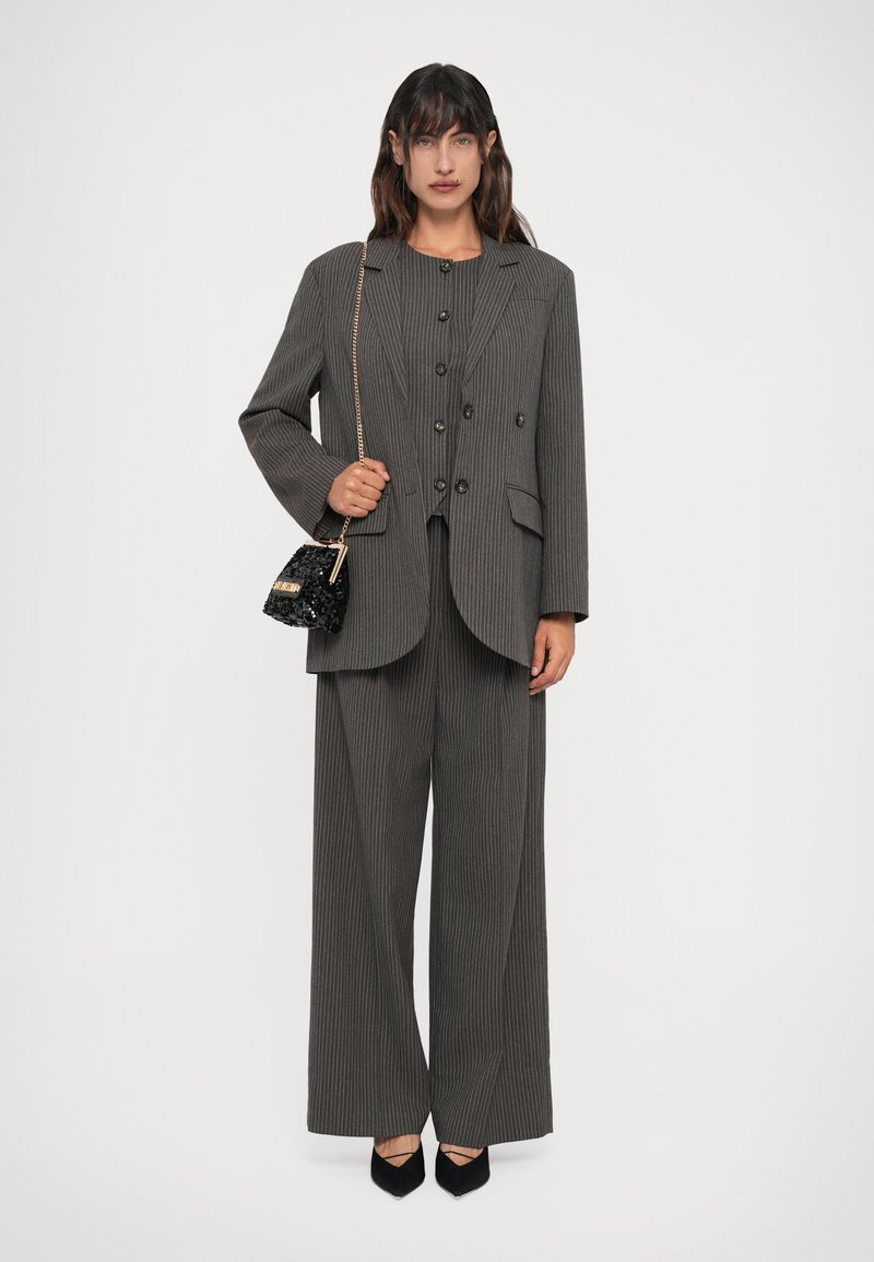 Donna in piedi con un pantalone grigio a righe sottili, che tiene una piccola borsa nera di paillettes con una catena dorata, indossando scarpe nere a punta.