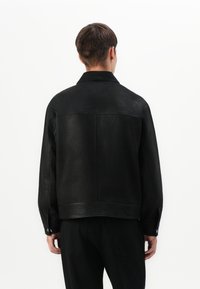 Man in een zwart leren jack en zwarte broek, staand met zijn rug naar de camera tegen een witte achtergrond.