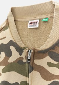 Camo ritsvest met een zachte beige stof en groene en bruine patronen. Ribgebreide kraag en een merkteken aan de hals.