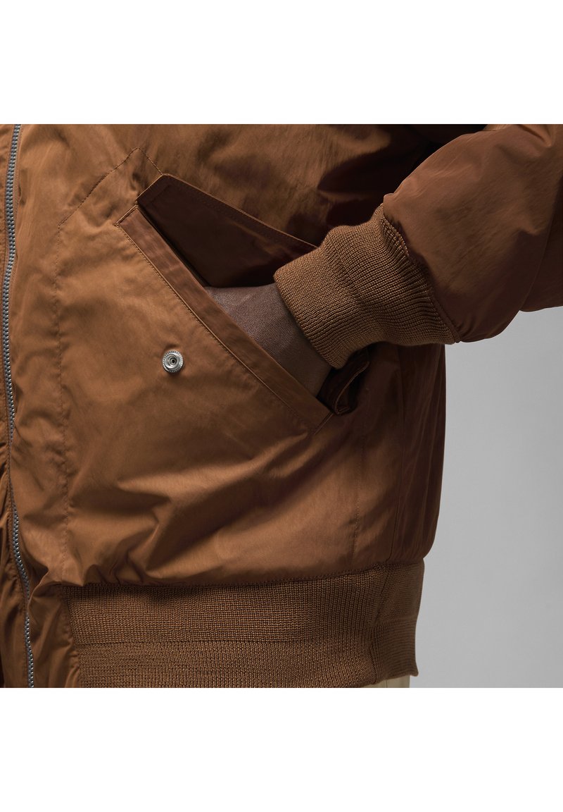 Braune Bomberjacke aus glattem, glänzendem Stoff, mit Rippbündchen und einer Druckknopf-Tasche auf der Vorderseite für sicheren Stauraum.