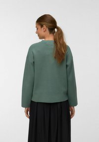 Ženska z rjavim ponytailom, oblečena v ohlapen zeleni pulover z dolgimi rokavi in črno nabrano krilo, stoji ob ozadju z enostavno belo barvo.