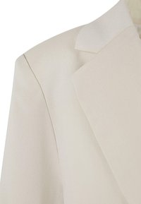 Blazer beige su misura con collo strutturato, revers affilati e texture del tessuto liscia. Dettagli delle cuciture evidenti sulla spalla.