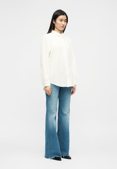 Γυναίκα που στέκεται φορώντας άνετο λευκό πουκάμισο με κουμπιά, μπλε τζιν παντελόνι φαρδιάς γραμμής με ξεφτισμένα τελειώματα και μαύρα μυτερά παπούτσια, μπροστά σε απλό φόντο.