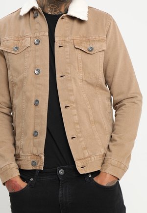 Homme portant une veste beige boutonnée avec un col en peau lainée blanche, une chemise noire en dessous et un jean noir, les mains dans les poches.
