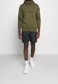 Sweat à capuche vert olive avec logo, en mélange de coton, poche kangourou à l'avant. Shorts noirs, légers, d'une longueur au-dessus du genou. Baskets blanches à lacets.