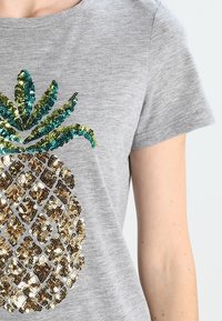 Camiseta gris de manga corta con un diseño de piña hecho de lentejuelas doradas y azul turquesa, con una textura suave y un cuello redondo.
