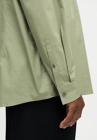 Δεξί χέρι ατόμου που φορά ανοιχτοπράσινο μακρυμάνικο πουκάμισο με κουμπωμένο μανσέτα και μαύρο παντελόνι, το χέρι χαλαρό στο πλάι.