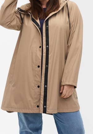 Femme portant un imperméable beige à capuche avec une bande de boutons noirs, associé à un jean bleu, debout sur un fond clair uni.