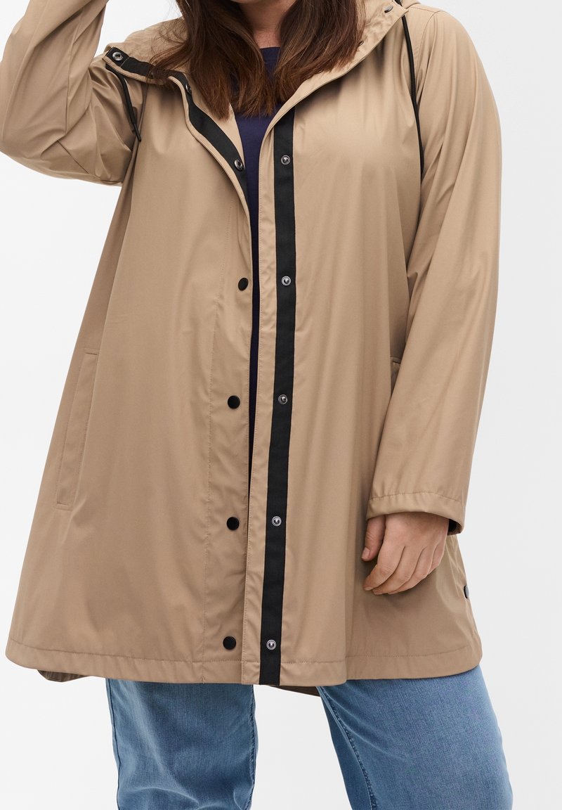 Femme portant un imperméable beige à capuche avec une bande de boutons noirs, associé à un jean bleu, debout sur un fond clair uni.