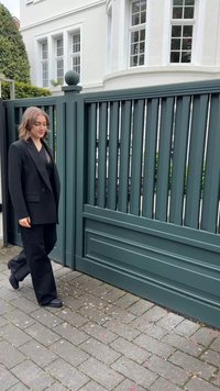 Blazer nero oversize e pantaloni a gamba larga indossati da una persona, che cammina su un sentiero di pietra accanto a un cancello verde in legno con listelli verticali.