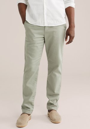 Homme portant un pantalon vert clair, une chemise blanche à manches longues et des chaussures beige à enfiler, debout avec une main dans la poche.