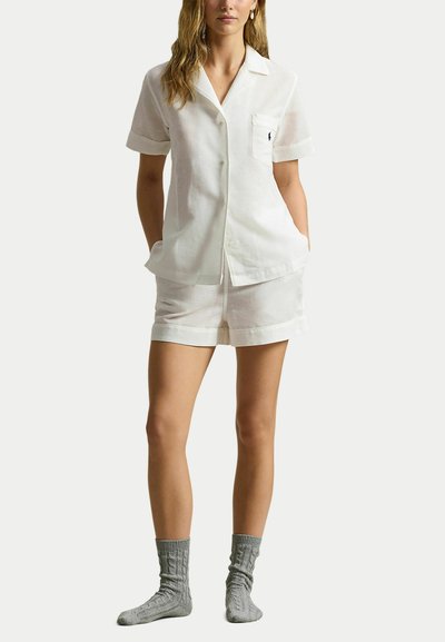 Vrouw draagt een witte korte mouwen blouse met knopen en een zak, gecombineerd met bijpassende korte broek, staat met handen in de zakken en draagt grijze gebreide sokken.