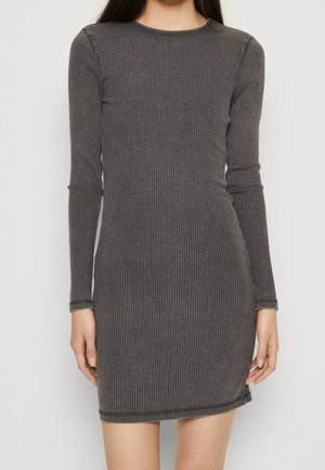 Kobieta ubrana w dopasowaną, długorękawową, prążkowaną, ciemnoszarą dzianinową sukienkę sięgającą połowy uda na jednolitym jasnym tle.